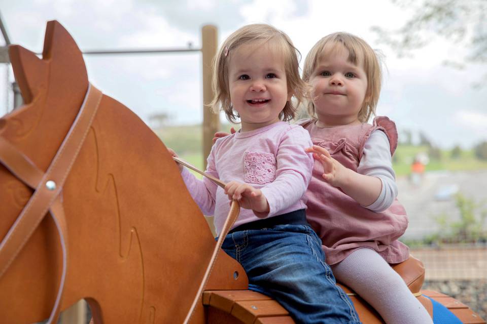 zwei Kleinkinder sitzen auf Spielplatz auf einem Holzpferd