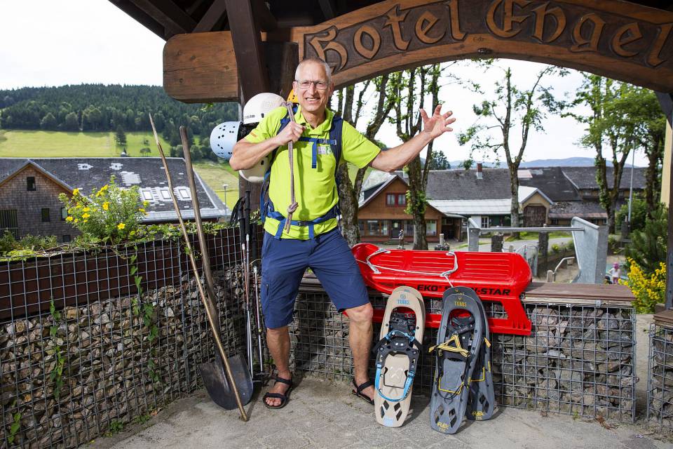 Mann steht vor Hotel Engel Schild mit Helmen, Schlitten und Schneeschuhen in kurzer Hose.