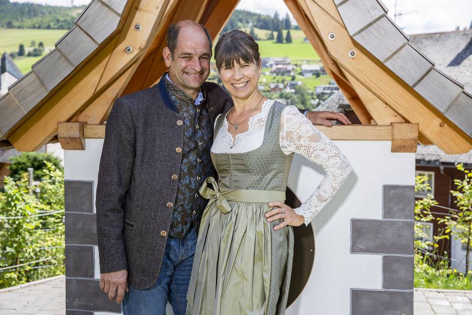 Gastgeber stehen vor einer Hütte in Tracht gekleidet und lächeln.