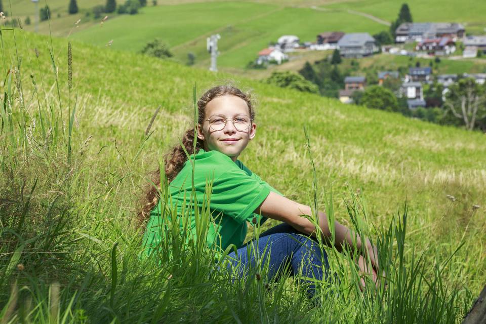 Mädchen sitzt entspannt im Gras mit Blick auf ein Dorf im Hintergrund