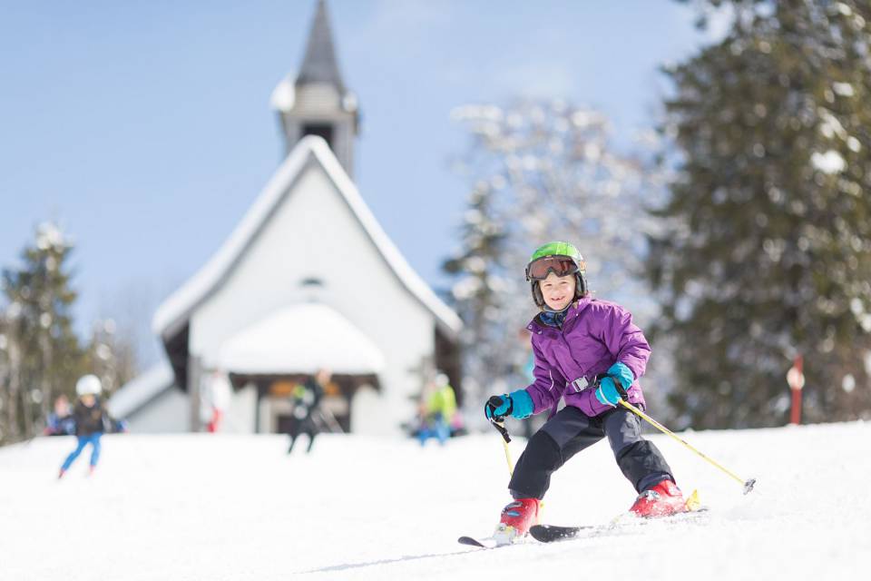 Kind mit bunter Winterkleidung fährt vor einer kleinen Kapelle Sk