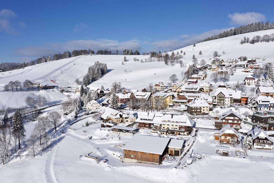 Luftaufnahme eines Bergdorfes mit Skihängen im Hintergrund.