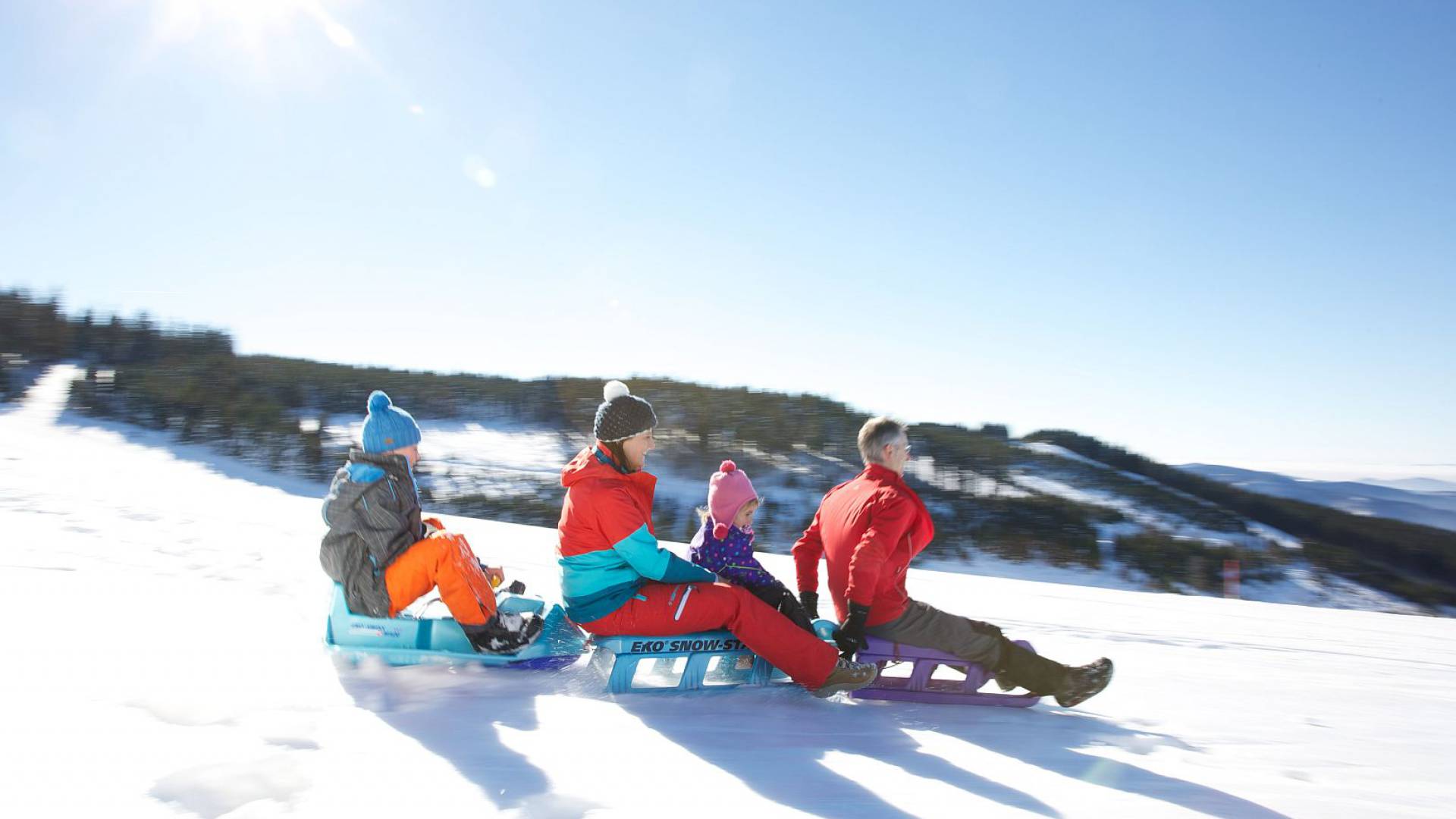 Familie mit zwei Kindern fährt auf drei Schlitten im Schnee den Berg herunter.