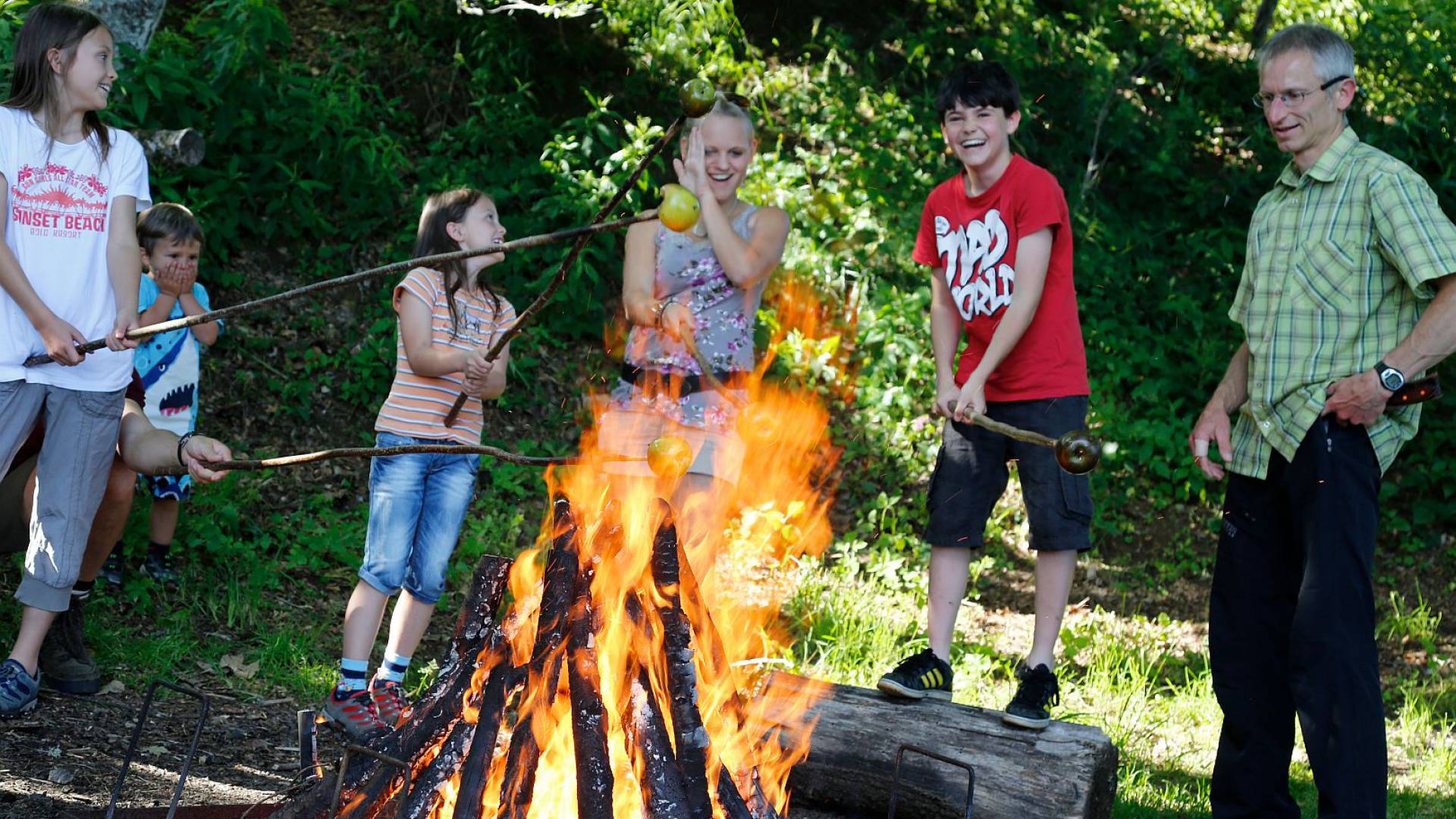 Einige Kinder und ein Mann stehen lachend am Lagerfeuer und braten Äpfel an