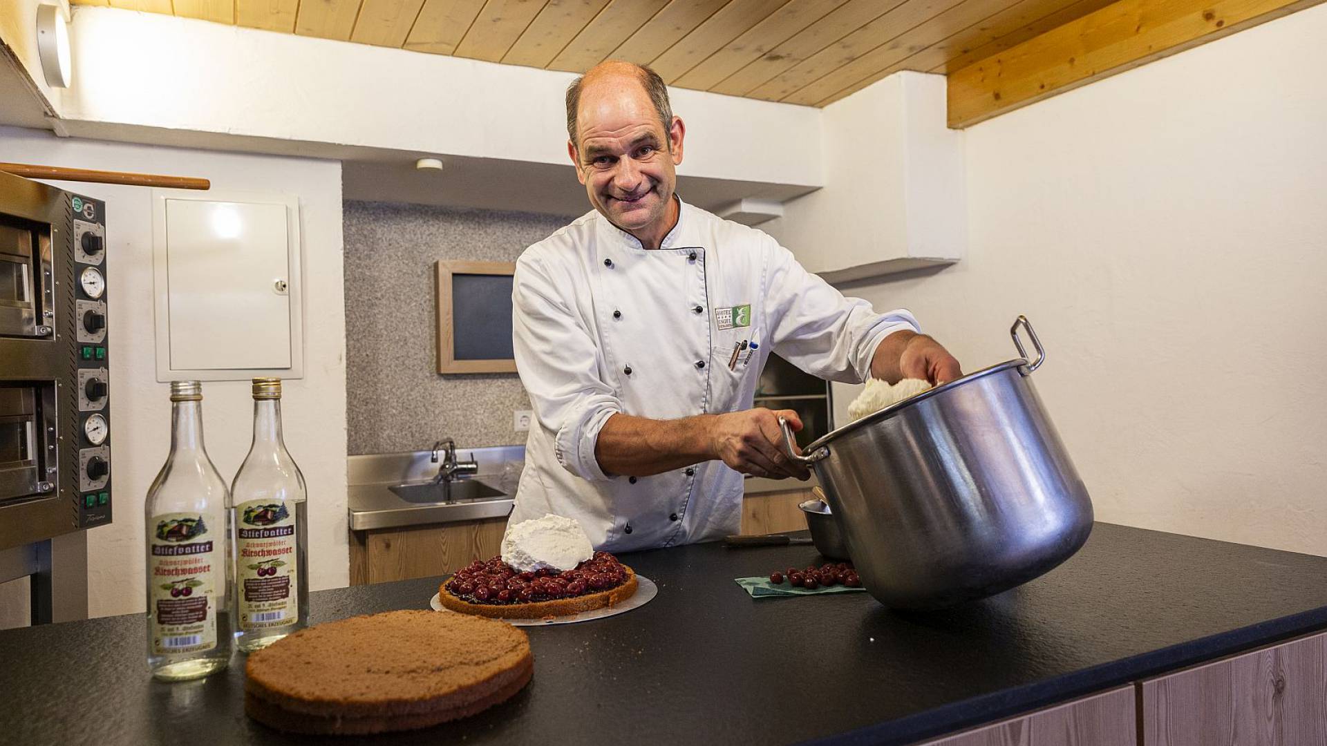 Koch verteilt Sahne auf einen Kuchenboden der mit Kirschen belegt ist, in einer Küche.