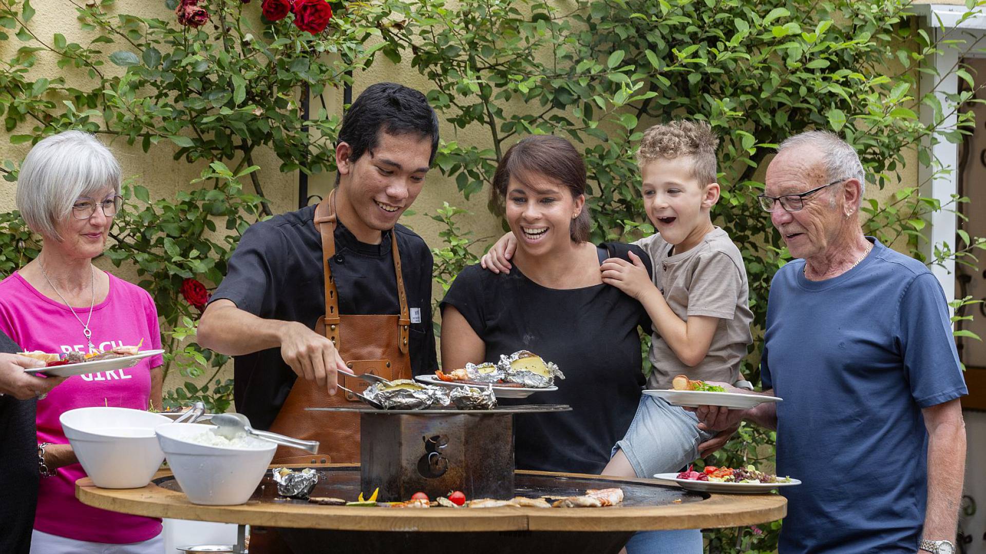 Koch grillt mit Familie an einer runden Feuerstelle im Garten.