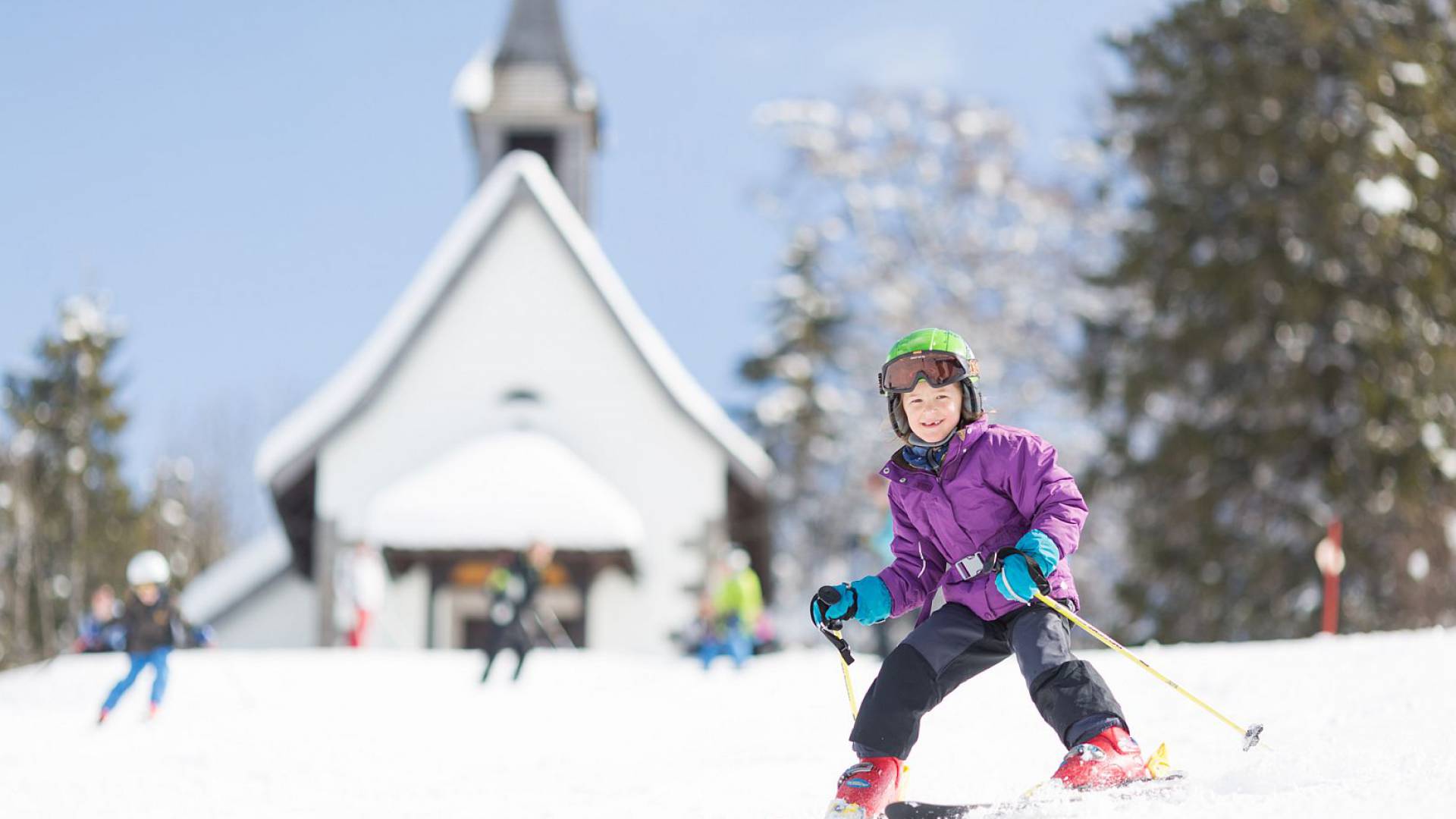 Kind mit bunter Winterkleidung fährt vor einer kleinen Kapelle Sk