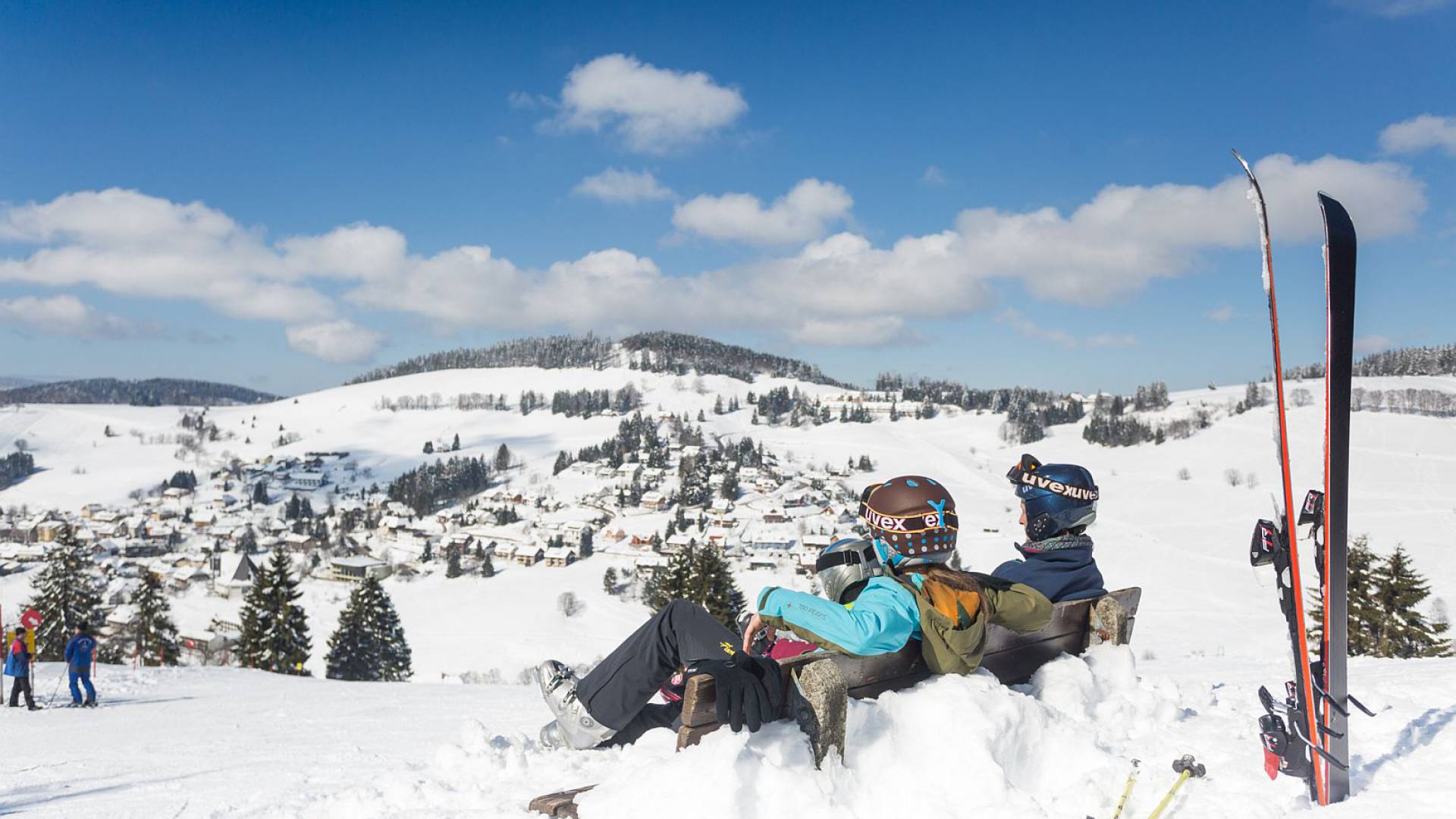 Skiausrüstung und Rucksack auf einem schneebedeckten Berghang abgelegt, Familie sitzt auf Bank.