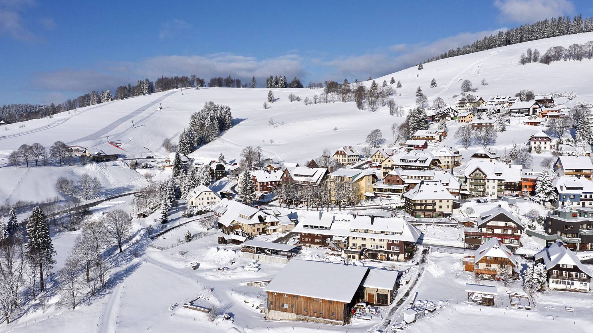 Luftaufnahme eines Bergdorfes mit Skihängen im Hintergrund.