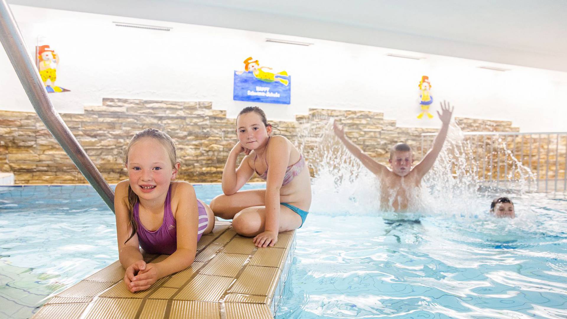 Mädchen sitzen am Poolrand, während Jungs im Wasser planschen